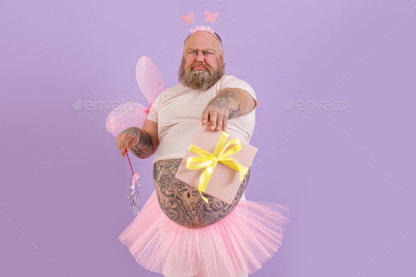 Obese man wearing fairy costume holds magic stick and gift box on purple background