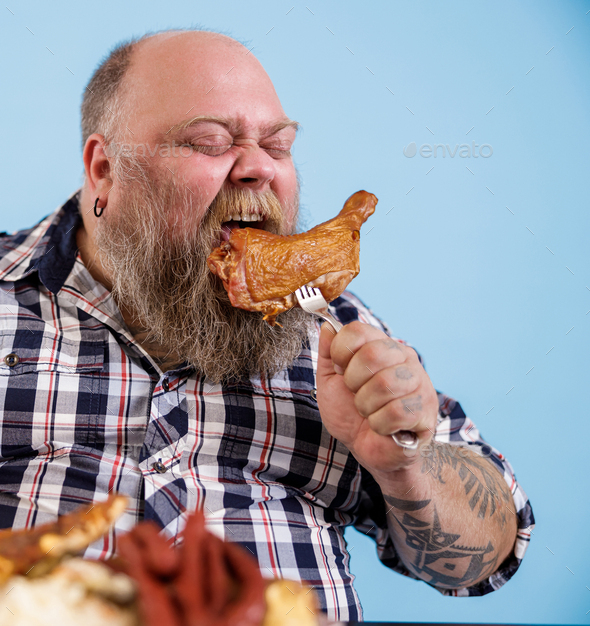 Bearded man with overweight bites piece of chicken leg on light blue ...