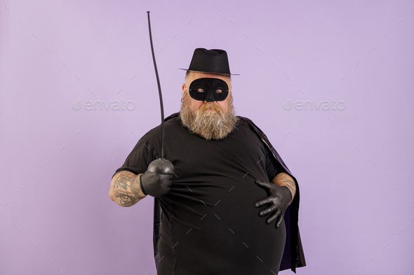 Mature man with overweight in Zorro costume holds hand on large tummy on purple background