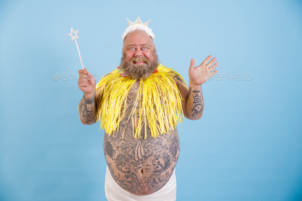 Joyful bearded plump man with crown, magic stick and yellow cape on light blue background
