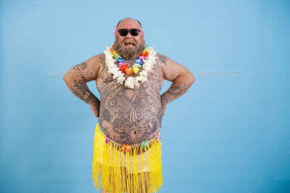Happy man with overweight and tattooed tummy in decorative grass skirt on light blue background