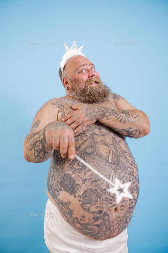 Plus size man with toy crown and magic stick poses on light blue background