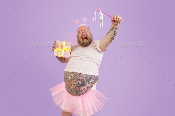 Excited man with overweight in fairy costume holds magic stick and present on purple background