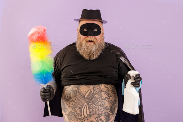 Emotional plump man in hero suit holds brush and spray bottle on purple background