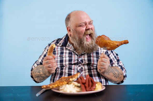 Hungry man with overweight eats chicken leg at table with fat food on blue  background Stock Photo by astakhovyaroslav