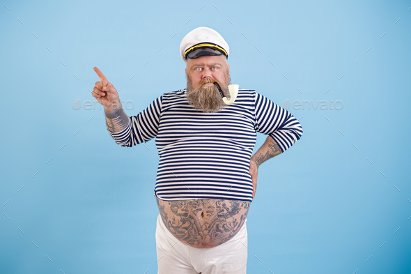 Plus size sailor man with tattoos and smoking pipe points aside by finger on light blue background