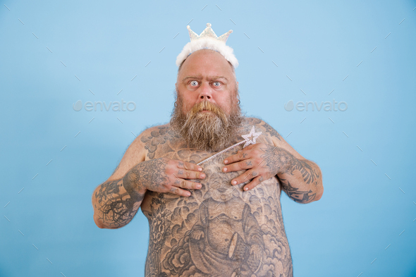 Funny obese man with toy crown and magic stick covers breasts on light blue background