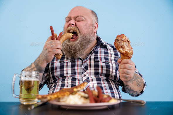 Hungry plus size man eats sausages at table with rich food and mug of beer