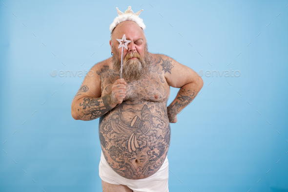Tattooed obese man with crown looks through star on magic stick on blue background