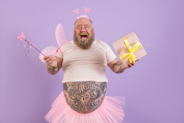 Joyful obese man in fairy costume holds gift box and magic stick on purple background