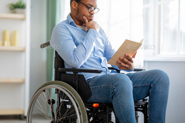 Smart impaired black man in wheelchair reading book, studying or working from home, selective focus