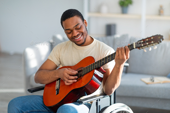 Positive paraplegic black man in wheelchair playing guitar, enjoying music at home - Stock Photo - Images