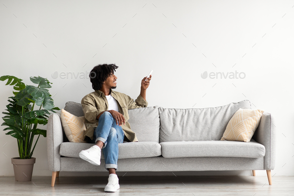 Happy black guy relaxing on couch, opening air conditioner with remote controller