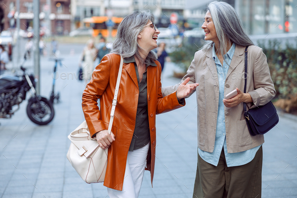 Positive senior women friends talk walking along modern city street ...