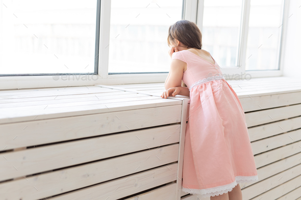 Charming little girl in a lush peach princess dress looks out the window of her room. The concept of