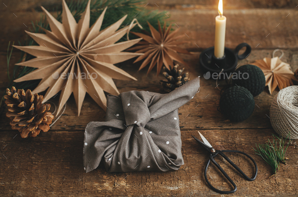 Stylish christmas gift wrapped in fabric on rustic table with scissors,  paper star, candle.Furoshiki Stock Photo by Sonyachny