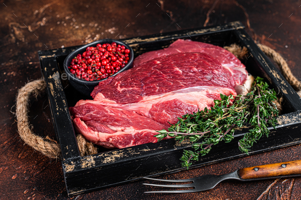 Fresh Raw Chuck eye roll beef steak in a wooden tray with herbs. Dark background. Top view