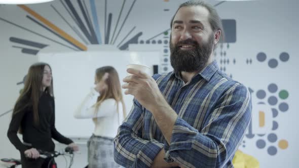 Positive Beardie in Checkered Shirt Drinking Coffee Looks Back at His Female Colleagues alt