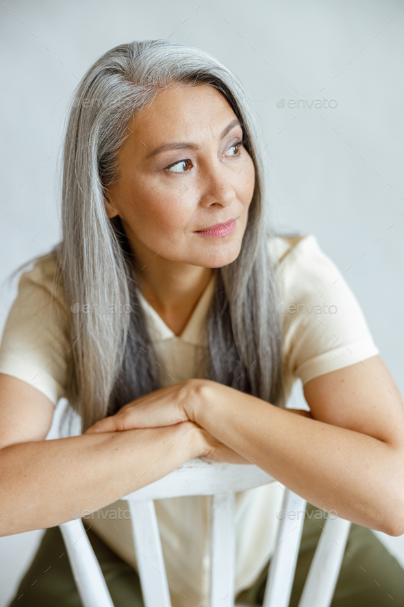 Pretty middle aged Asian lady with straight grey hair sits backwards on chair in studio