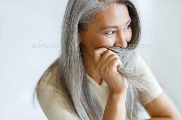 Middle aged Asian woman hides face with grey hair sitting on light background