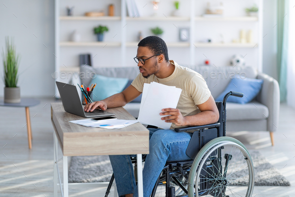 Paraplegic black guy in wheelchair with documents using laptop to work online from home - Stock Photo - Images