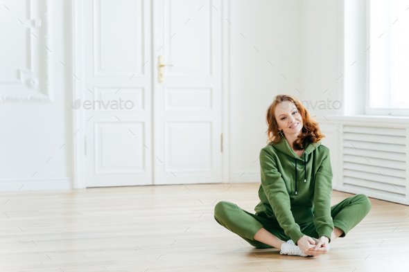 Delighted redhead female sits on lotus pose, wears tracksuit, being in good body shape