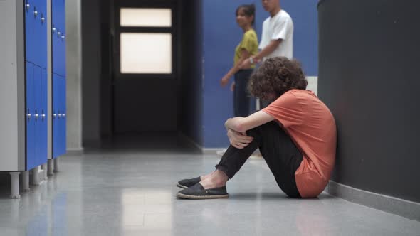 Sad Teenager Sitting Alone on Floor Victim of Bullying While Classmates Ignore Him alt