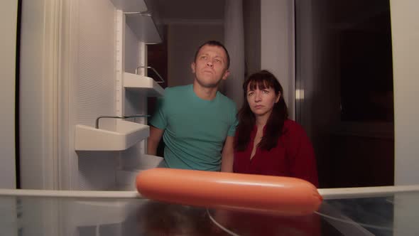 Hungry Man and Woman Open the Refrigerator at Night and Take a Sausage