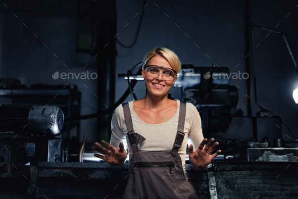 Happy young industrial woman with dirty hands looking at camera indoors in metal workshop.
