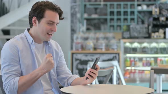 Creative Man Celebrating on Smartphone in Cafe alt