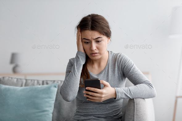 Sad shocking european young lady suffering from depression sits on sofa read message