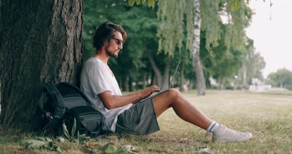 Bearded Man Is Working with Laptop at the Park Under the Tree. Happy Freelancer Is Sitting and Using alt