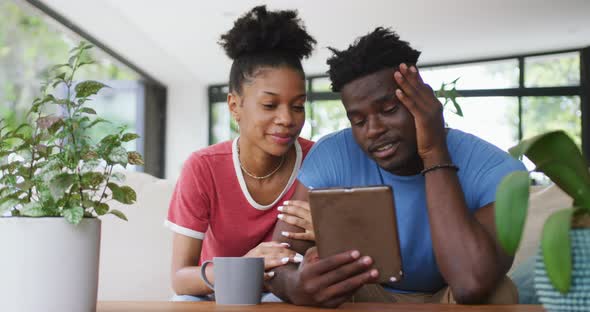 Video of happy african american couple sitting on sofa and using tablet alt