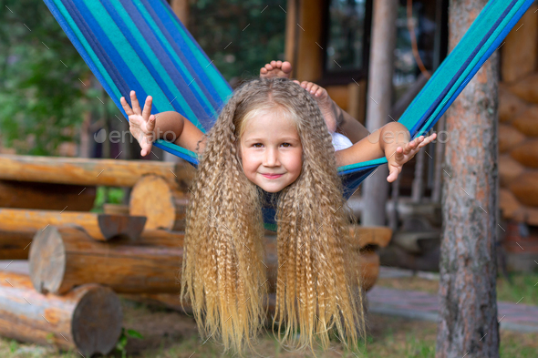 Little happy girl with long blond curly hair sways spread arms on a blue-green hammock.