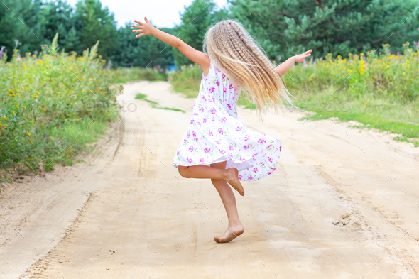 A little girl with long blond curly hair is dancing, spinning on a forest road