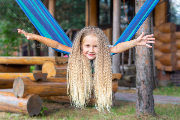 Little happy girl with long blond curly hair sways spread arms on a blue hammock. Summer carefree.