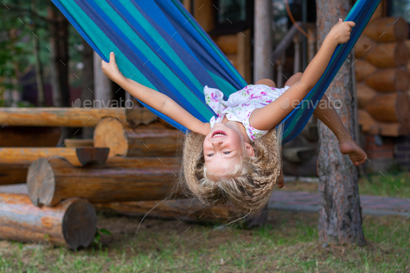 Little happy girl with long blond curly hair sways upside down on a blue-green hammock.