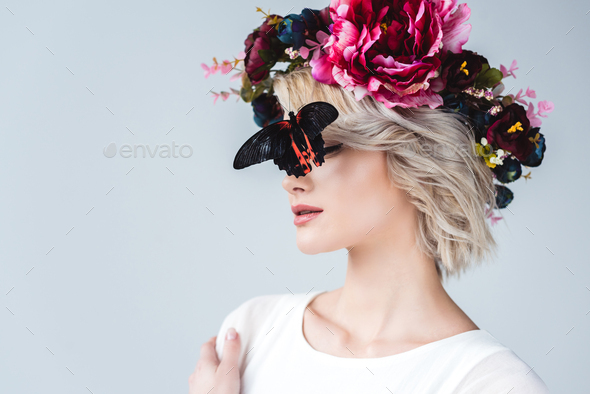 stylish girl posing in floral wreath with beautiful alive butterfly on face, isolated on grey