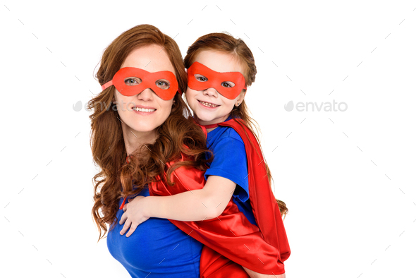 super mother piggybacking adorable daughter in mask and cloak and smiling at camera isolated on