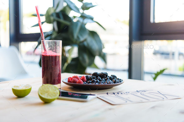 close up view of detox drink in glass, lime pieces, fresh berries and smartphone on table