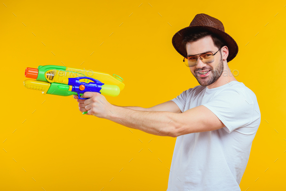 stylish young man holding water gun and smiling at camera isolated on yellow