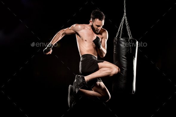 shortless boxer performing flying kick near punching bag isolated on black