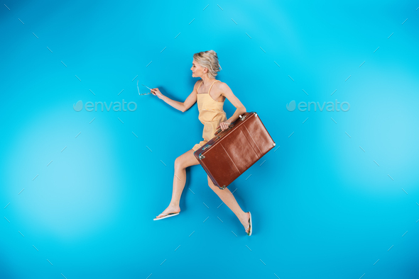 full length view of beautiful young woman in flip flops holding suitcase and running on blue