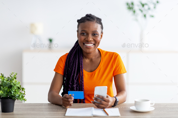 Happy black lady sitting at desk with cellphone and credit card, shopping on internet at home