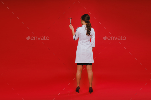 a beautiful doctor girl holds a reflex hammer and smiles at the camera isolated on a Red background