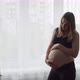 A Young Pregnant Woman Strokes Her Belly with Her Hands Near the Window