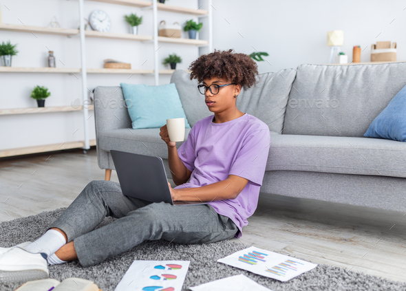 Black teenage student making educational project for online course, drinking coffee at home