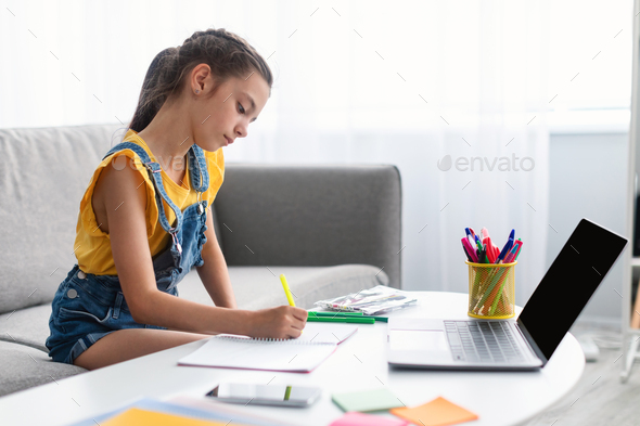 Girl sitting at desk, using laptop, writing in textbook