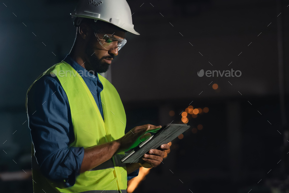 Young industrial man with protective wear using tablet indoors in metal workshop at night.
