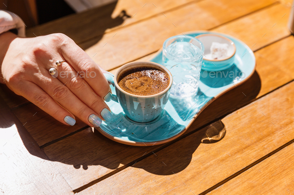 a cup of hot black turkish coffee in blue cup in women\'s hands served with glass of water and sugar